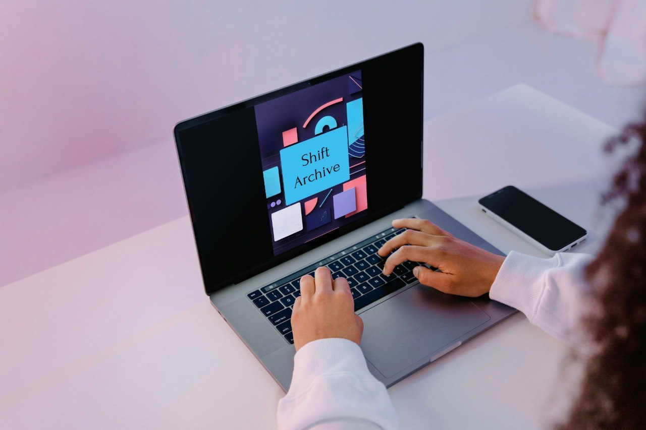 A person using a silver laptop on a white desk, with their hands on the keyboard and mouse. The laptop screen displays a colorful graphic with the text 'Shift Archive'.
